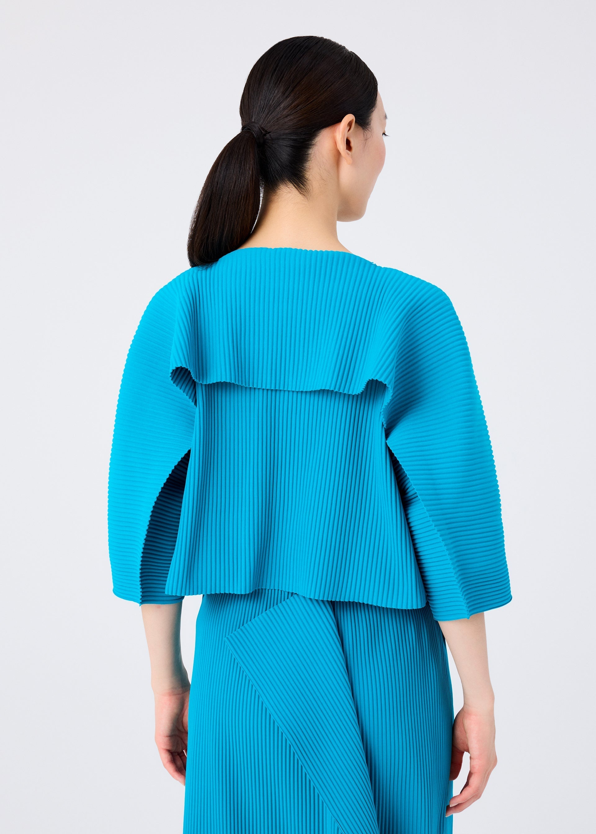 Zoomed view A woman with dark hair in a low ponytail is seen from behind, wearing the bright blue TRICK PLEATS top and skirt by me ISSEY MIYAKE, featuring fine knit pleats for a striking modern look.