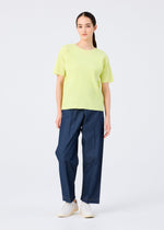 A person stands against a plain white background, wearing the me ISSEY MIYAKE FINE KNIT PLEATS COLOR 2 top in light yellow with subtle textured pleats, paired with loose blue pants and white shoes. Their long dark hair is tied back; expression is neutral.