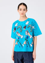 A young woman with dark hair in a low ponytail models the MAGIC FINE KNIT PLEATS T by me ISSEY MIYAKE, a bright blue short-sleeved top with a vibrant transfer print, against a plain white background.