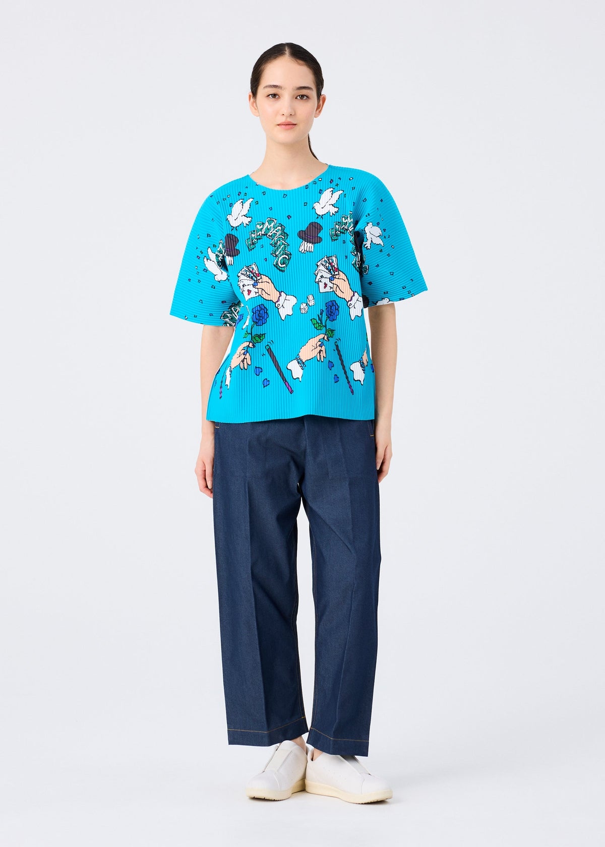 A person faces forward wearing the me ISSEY MIYAKE MAGIC FINE KNIT PLEATS T, a bright blue short-sleeved top with a colorful abstract floral pattern, paired with loose dark blue trousers and white slip-on shoes against a plain white background. view 6 of 7