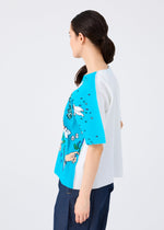 A woman with dark hair in a low ponytail wears the MAGIC FINE KNIT PLEATS T by me ISSEY MIYAKE, a loose, short-sleeved top with blue and white color blocking and playful transfer print patterns, seen from the side against a plain background.