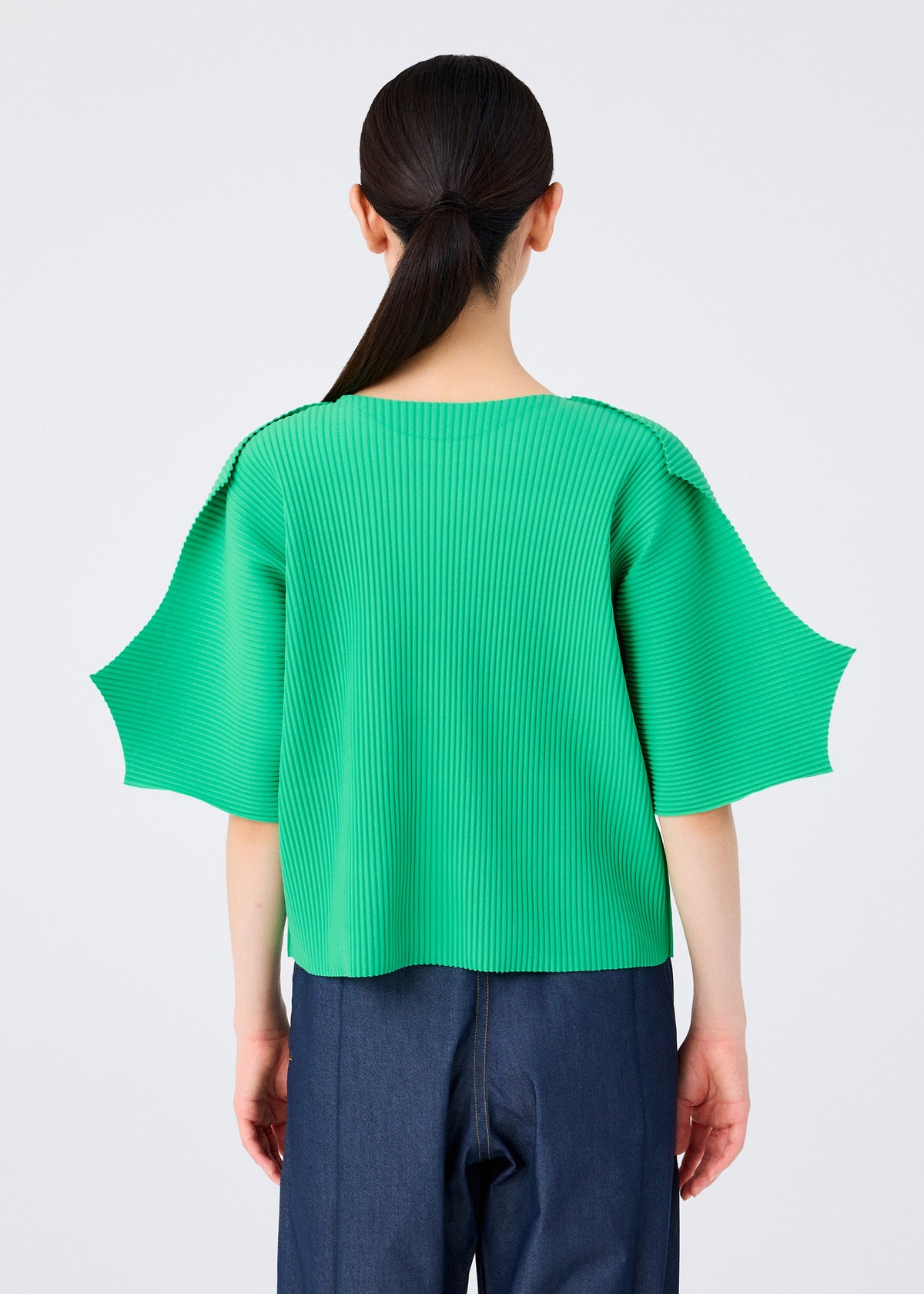 A person with long dark hair in a low ponytail is seen from behind, wearing the me ISSEY MIYAKE GIZA GIZA PLEATS bright green pleated knit top with wide short sleeves and dark blue jeans against a plain light background. view 7 of 8