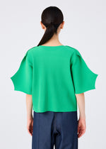 A person with long dark hair in a low ponytail is seen from behind, wearing the me ISSEY MIYAKE GIZA GIZA PLEATS bright green pleated knit top with wide short sleeves and dark blue jeans against a plain light background.