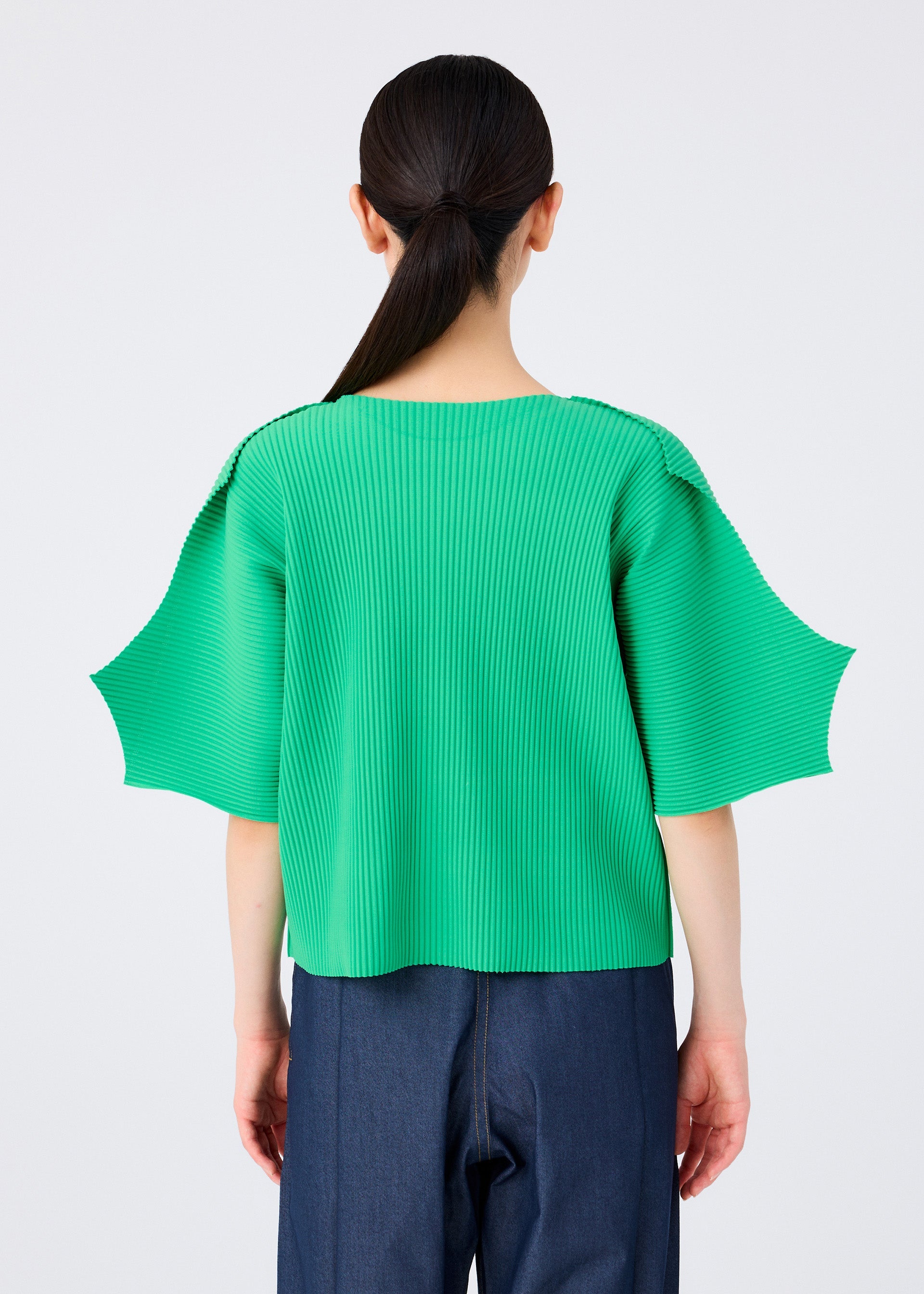 Zoomed view A person with long dark hair in a low ponytail is seen from behind, wearing the me ISSEY MIYAKE GIZA GIZA PLEATS bright green pleated knit top with wide short sleeves and dark blue jeans against a plain light background.