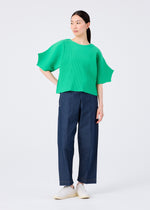 A person with long dark hair pulled back stands against a white background, wearing the me ISSEY MIYAKE GIZA GIZA PLEATS bright green pleated knit top with wide sleeves, dark blue straight-leg pants, and white shoes.
