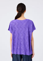 A person with long dark hair in a low ponytail is seen from behind, wearing the me ISSEY MIYAKE WAVE KNIT STRETCH PLEATS top and dark blue pants, set against a plain light background.