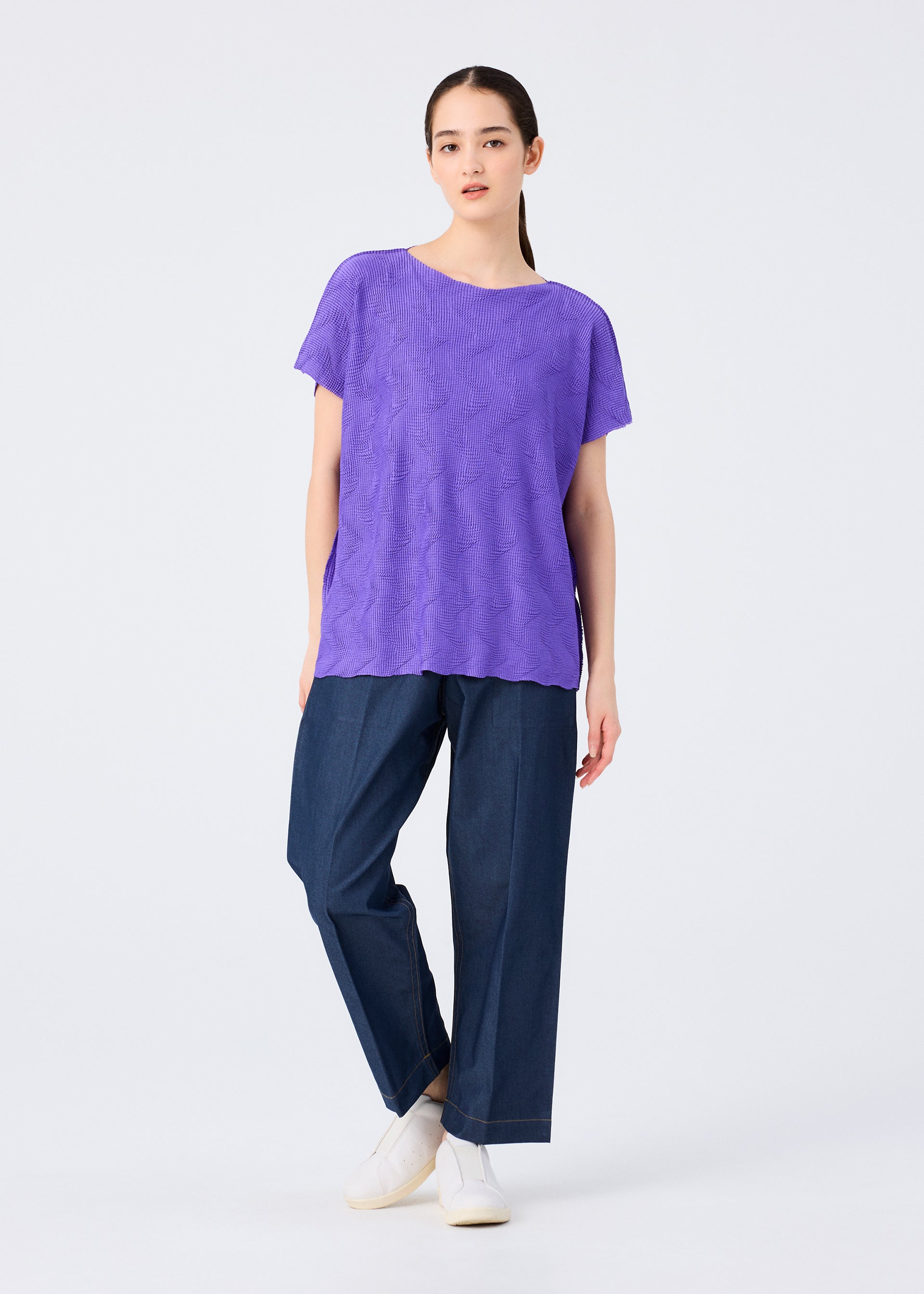 Zoomed view A woman poses against a white background wearing the me ISSEY MIYAKE WAVE KNIT STRETCH PLEATS purple top with wave patterns, wide-leg dark blue jeans, and white slip-ons; her straight hair is pulled back and one leg is slightly crossed.