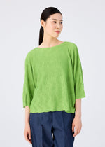 A person with straight, dark hair tied back is wearing the bright green WAVE KNIT STRETCH PLEATS top by me ISSEY MIYAKE with three-quarter sleeves and blue pants, standing against a plain light background.