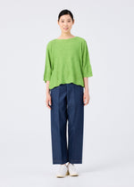 A person stands facing forward, smiling slightly. They wear the me ISSEY MIYAKE WAVE KNIT STRETCH PLEATS top in bright green with elbow-length sleeves, wide-leg blue jeans, and white shoes against a plain light background.