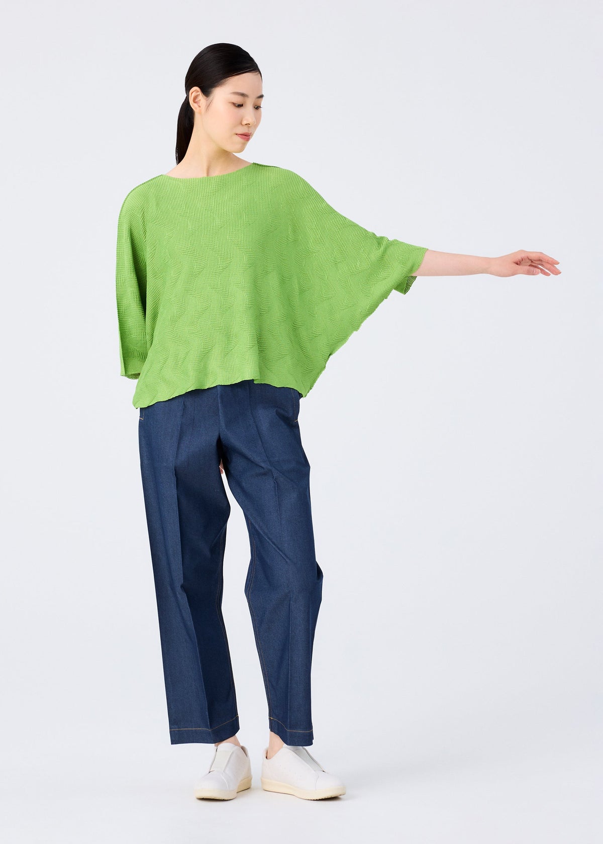 A person poses against a plain background in a me ISSEY MIYAKE WAVE KNIT STRETCH PLEATS bright green pleated knit top with short sleeves, wide dark blue jeans, and white shoes, one arm extended gracefully to the side. view 9 of 9