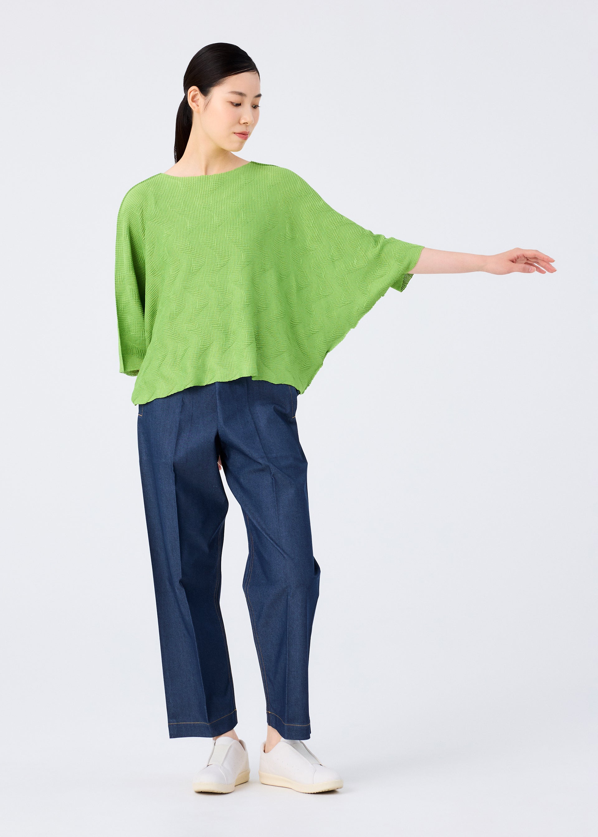 Zoomed view A person poses against a plain background in a me ISSEY MIYAKE WAVE KNIT STRETCH PLEATS bright green pleated knit top with short sleeves, wide dark blue jeans, and white shoes, one arm extended gracefully to the side.