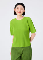 A person with straight, chin-length black hair is smiling in a bright green ROUND PLEATS short-sleeved top and matching pants by me ISSEY MIYAKE, standing against a plain light background.