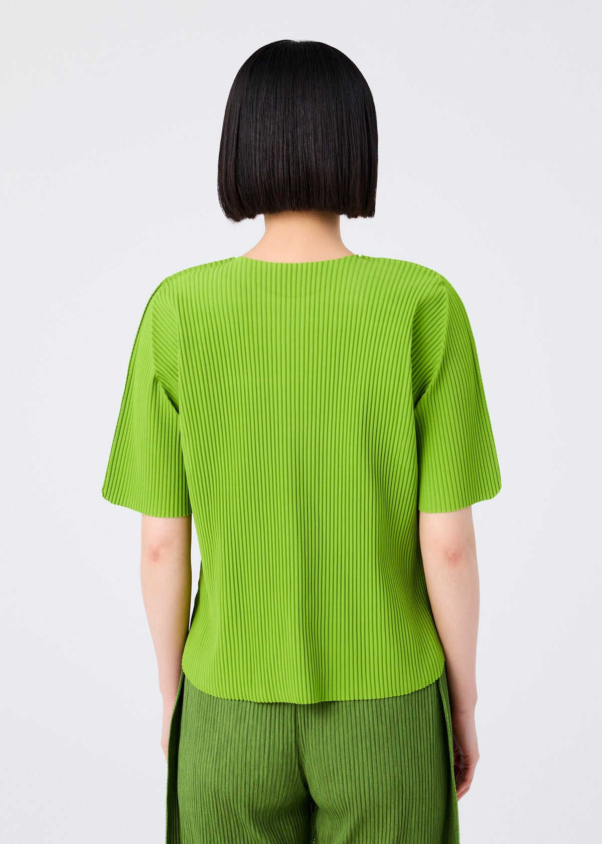 A person with a straight, chin-length black bob is seen from the back wearing the ROUND PLEATS short-sleeved knit top and matching pants in bright green by me ISSEY MIYAKE, set against a plain light background. view 6 of 7