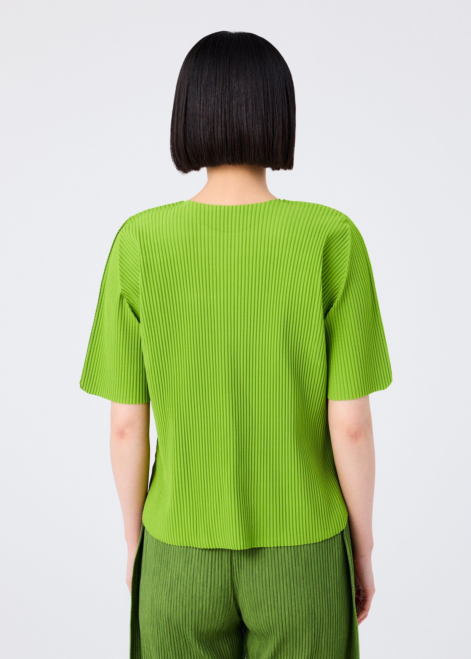 Zoomed view A person with a straight, chin-length black bob is seen from the back wearing the ROUND PLEATS short-sleeved knit top and matching pants in bright green by me ISSEY MIYAKE, set against a plain light background.
