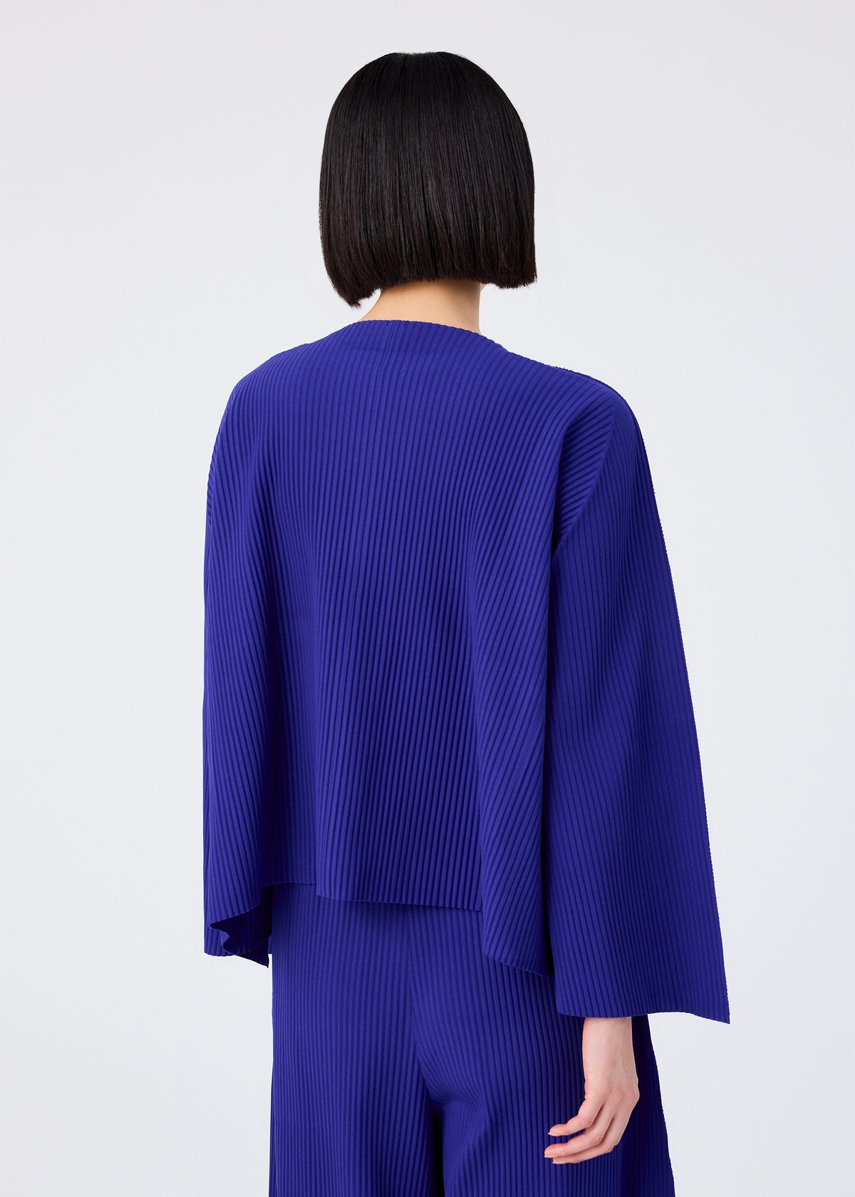 A person with straight, chin-length dark hair is shown from the back, wearing the me ISSEY MIYAKE FLARE PLEATS cardigan and pants in royal blue against a plain white background. view 8 of 9