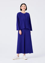 A person with short black hair is standing and smiling, wearing the FLARE PLEATS set from me ISSEY MIYAKE—loose pleated knit cardigan and matching wide-leg blue pants with white slip-on shoes against a plain white background.