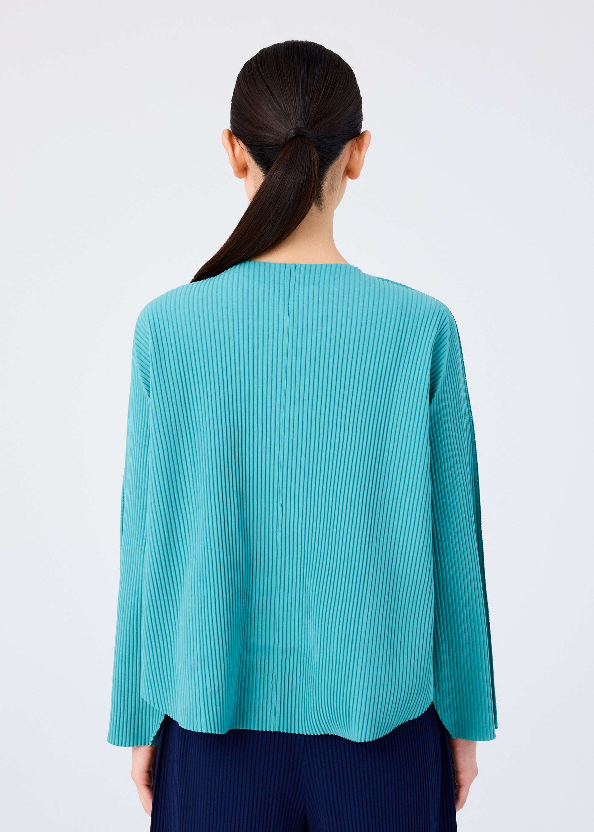 Zoomed view A person with long dark hair in a low ponytail is seen from the back, wearing the me ISSEY MIYAKE AURORA PLEATS teal knit cardigan and dark pants against a plain white background.