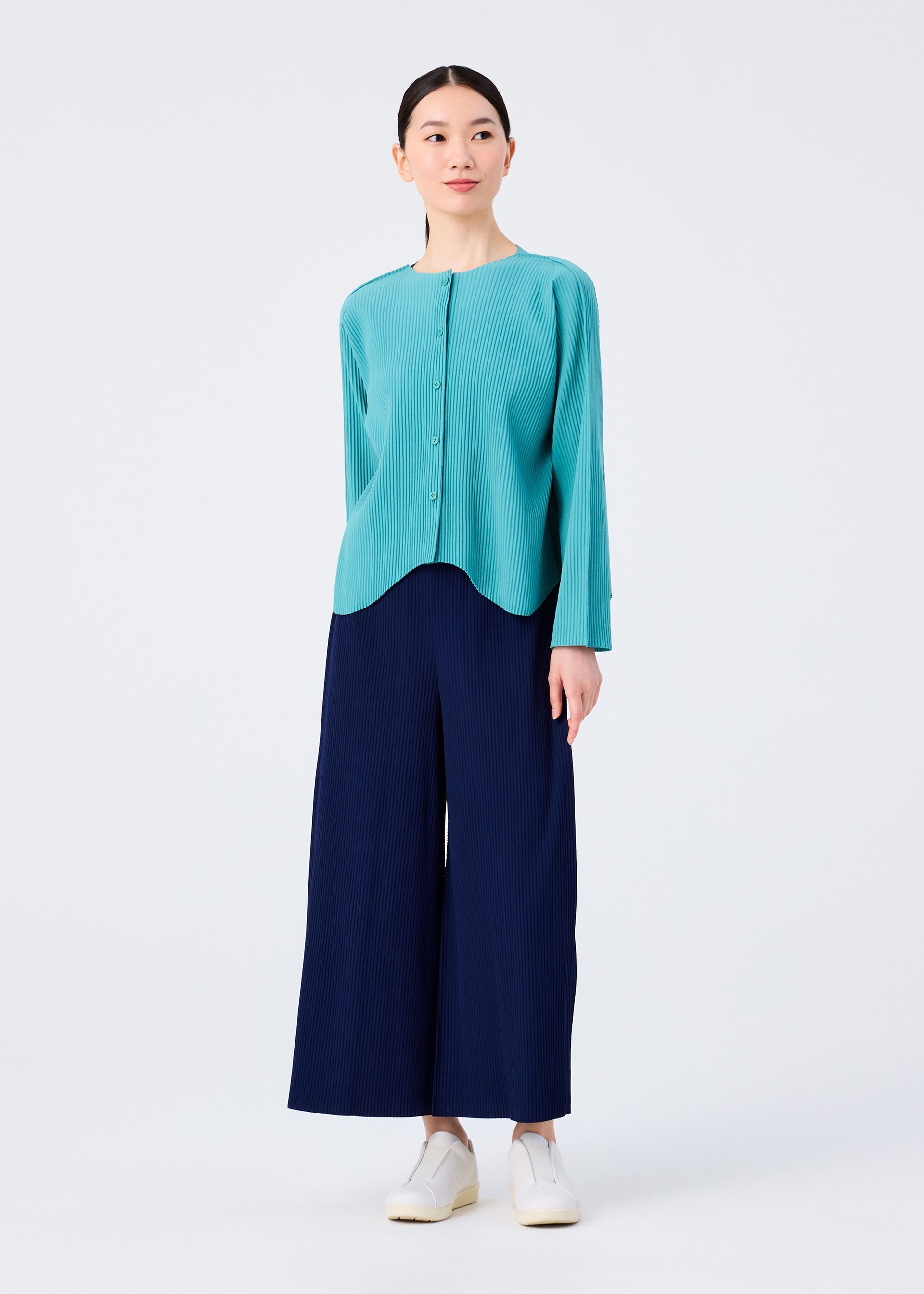Zoomed view A woman stands against a plain background wearing the me ISSEY MIYAKE AURORA PLEATS turquoise fine knit cardigan, paired with wide-leg navy pants and white slip-on shoes. Her straight black hair is pulled back as she gazes slightly to the side.