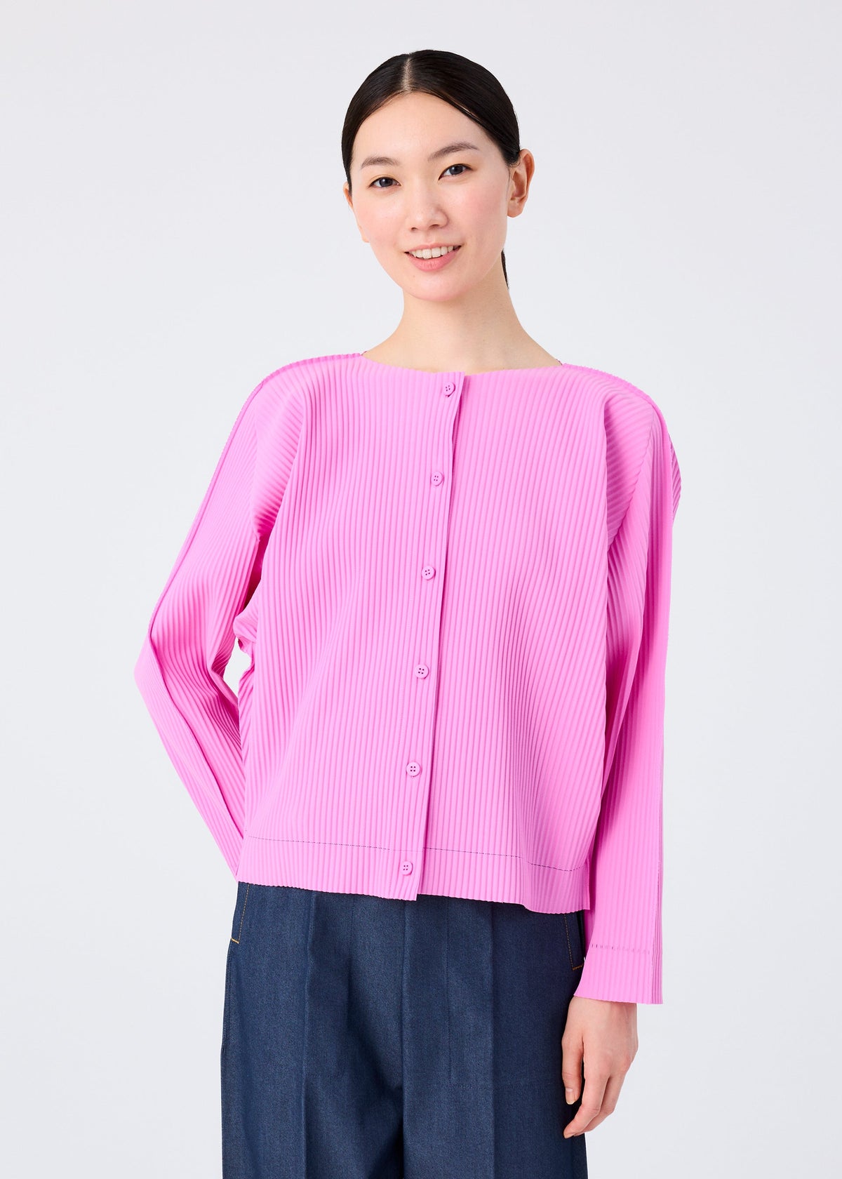 A person with dark hair tied back smiles, wearing the me ISSEY MIYAKE FINE KNIT PLEATS COLOR 2 light pink pleated cardigan and dark blue pants, standing against a plain white background. view 6 of 8