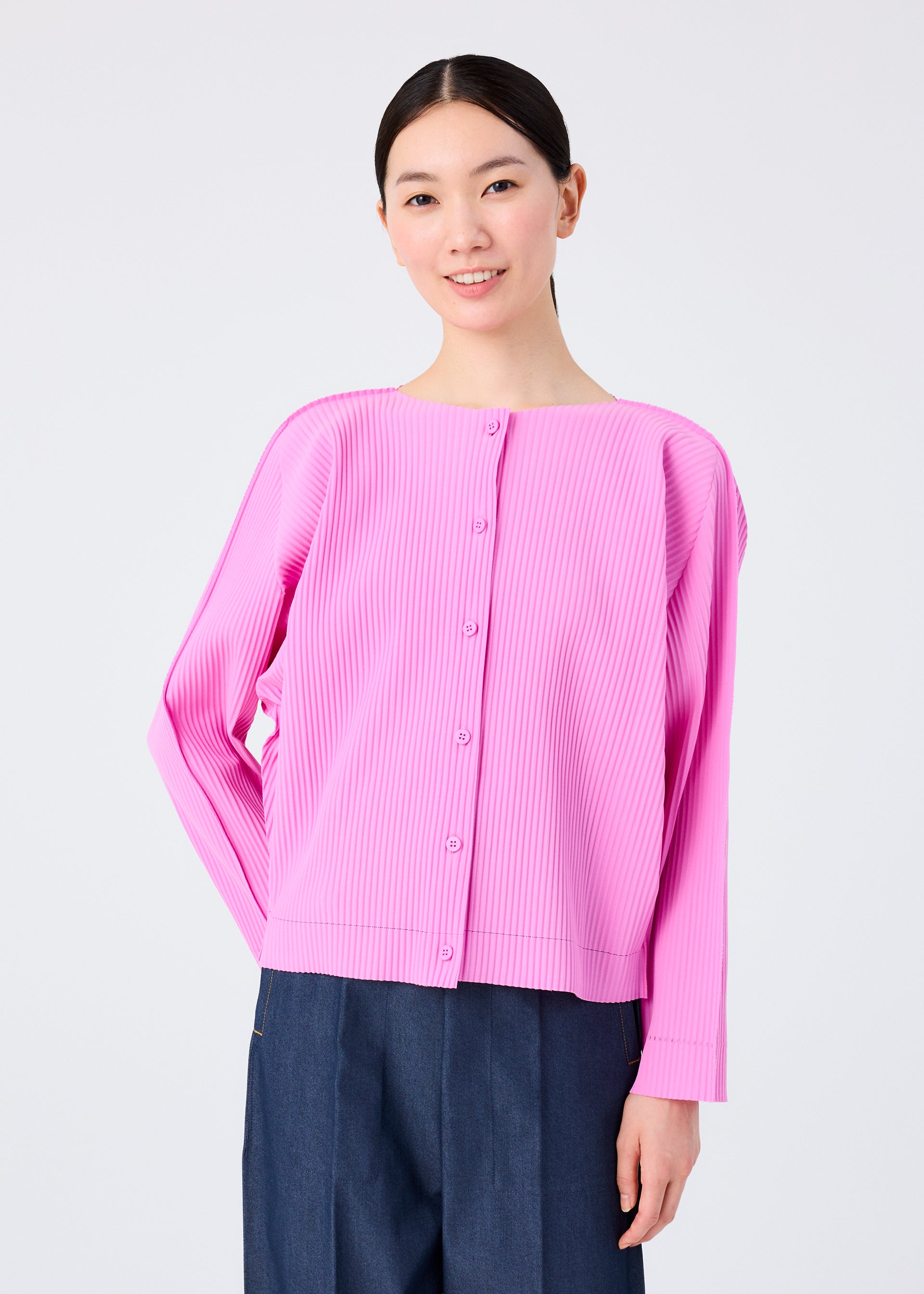 Zoomed view A person with dark hair tied back smiles, wearing the me ISSEY MIYAKE FINE KNIT PLEATS COLOR 2 light pink pleated cardigan and dark blue pants, standing against a plain white background.