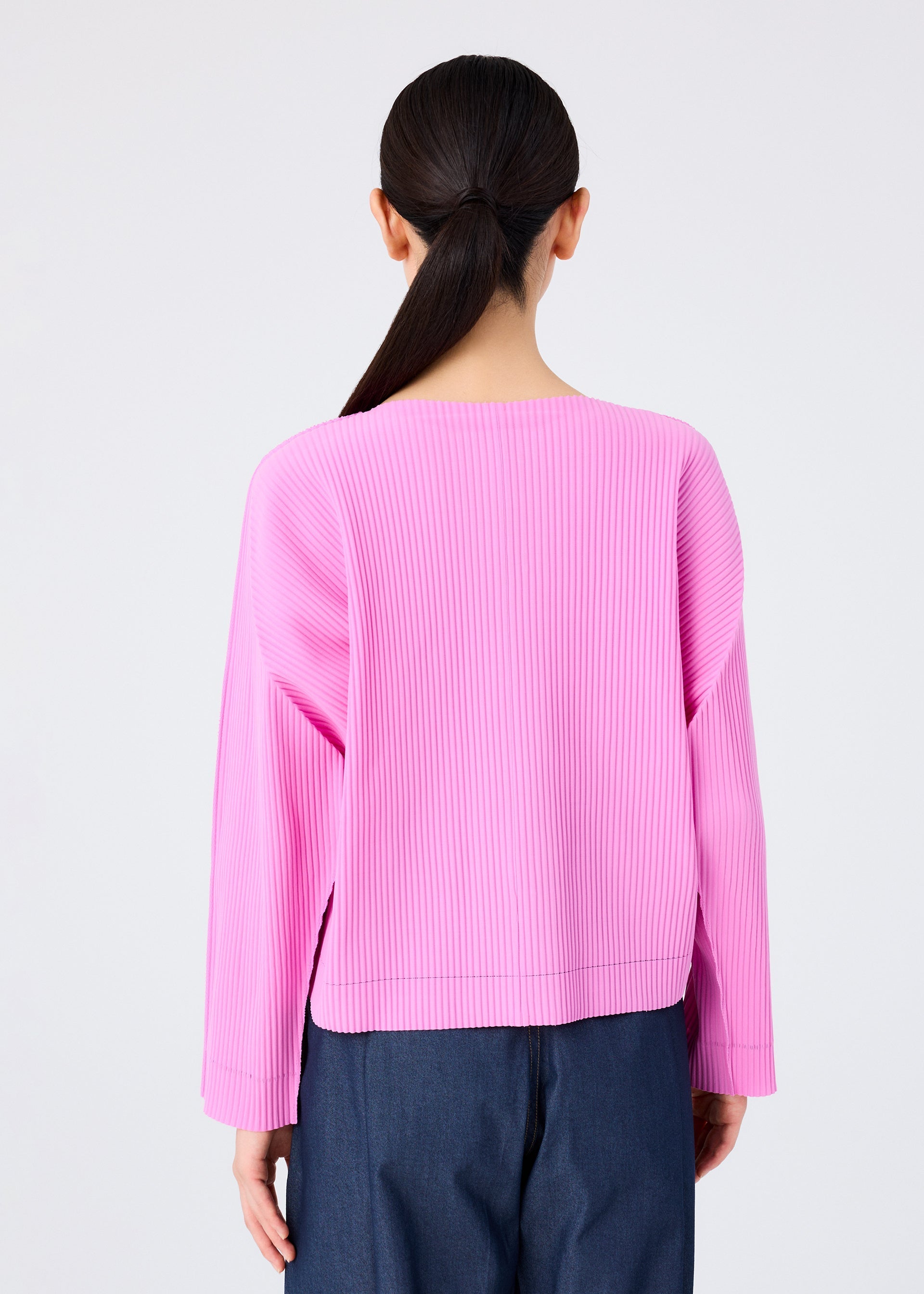 Zoomed view A person with long dark hair in a low ponytail is seen from the back, wearing the me ISSEY MIYAKE FINE KNIT PLEATS COLOR 2 cardigan in bright pink and dark blue pants, set against a plain light background.