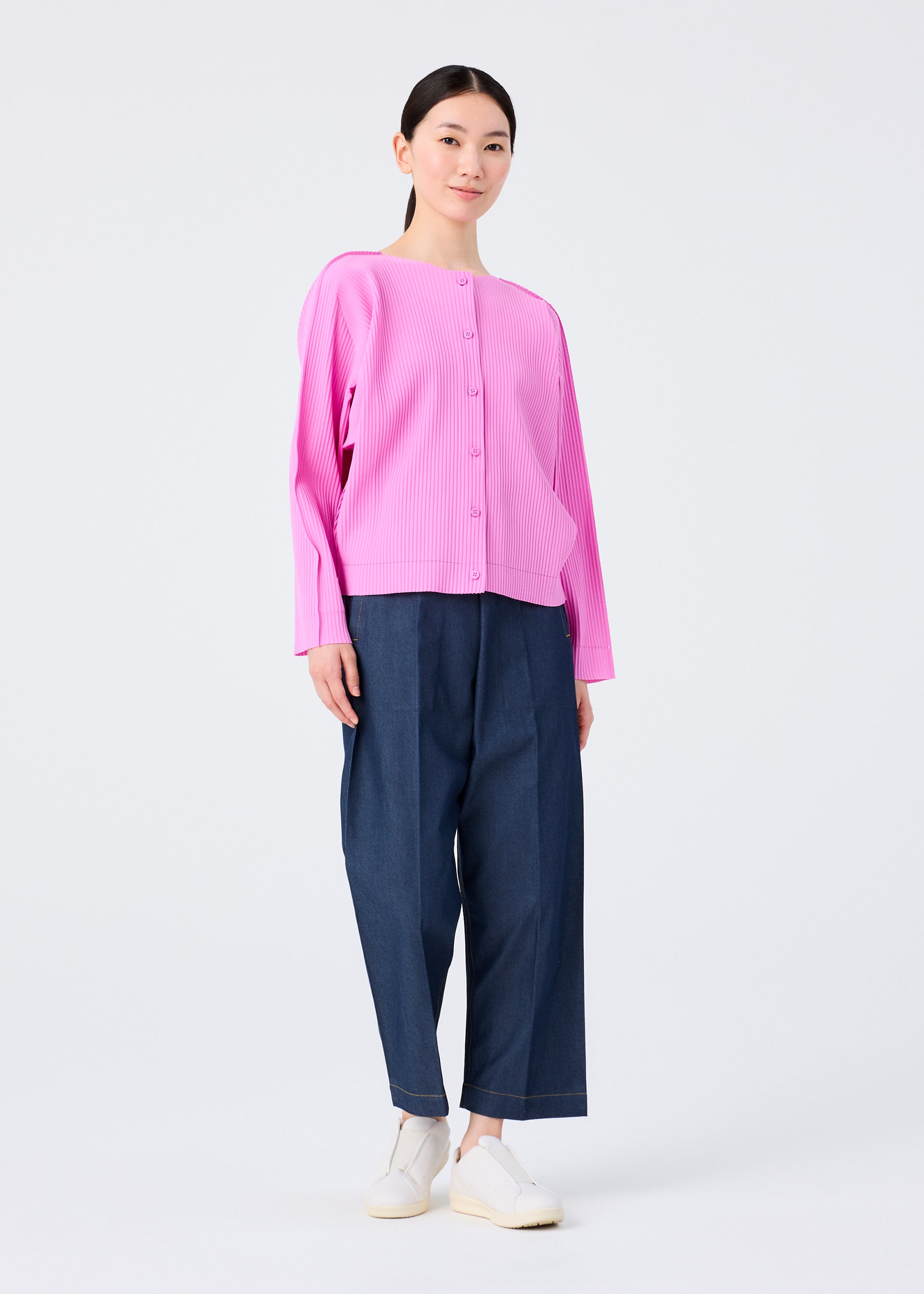 Zoomed view A woman with straight dark hair tied back and a neutral expression wears the me ISSEY MIYAKE FINE KNIT PLEATS COLOR 2—a bright pink, long-sleeved fine knit cardigan—paired with loose dark blue pants and white slip-on shoes against a plain background.