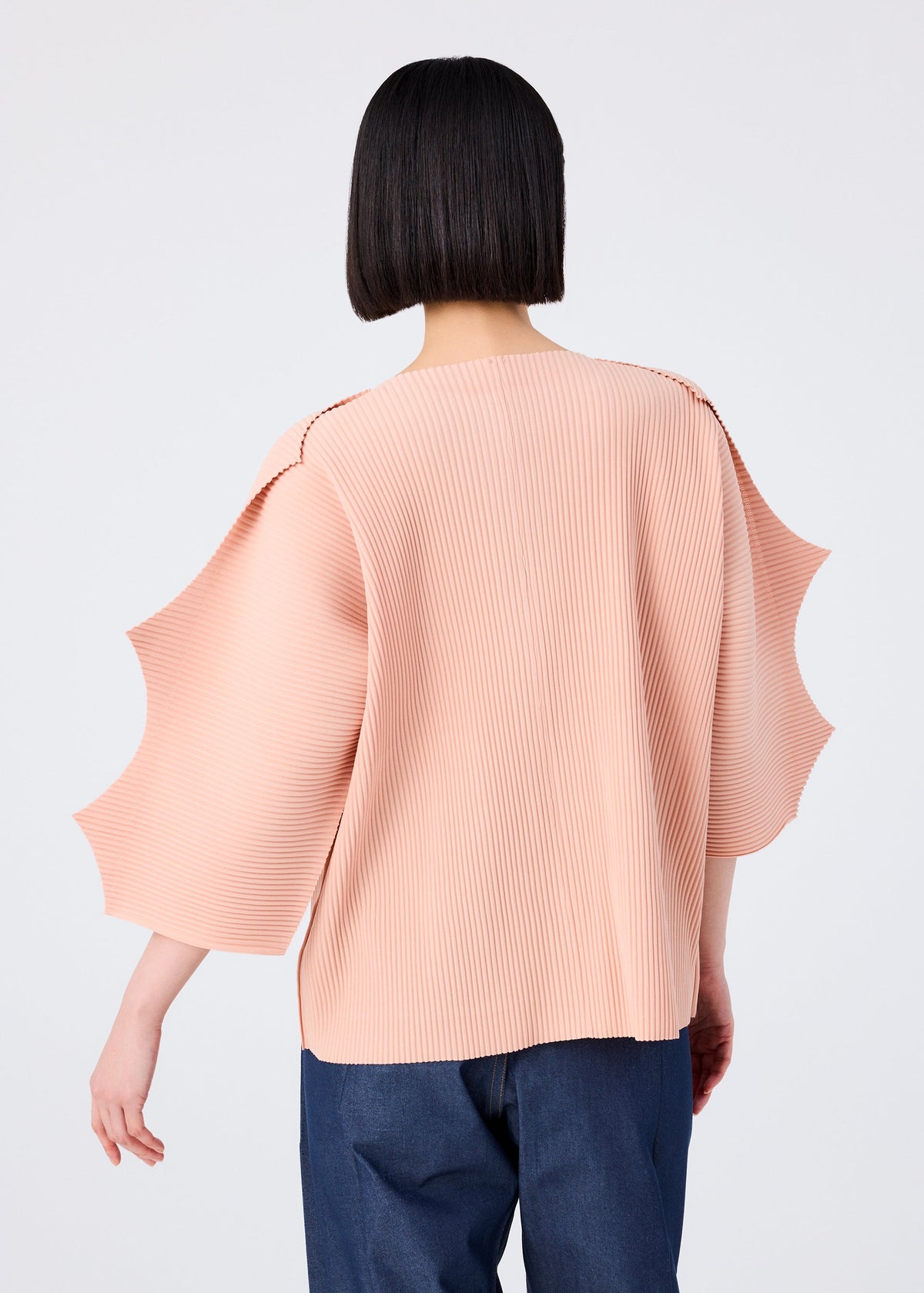 A person with a straight bob haircut is seen from behind, wearing the me ISSEY MIYAKE GIZA GIZA PLEATS pale pink pleated knit cardigan with sculpted sleeves and dark blue pants against a plain light background. view 8 of 10