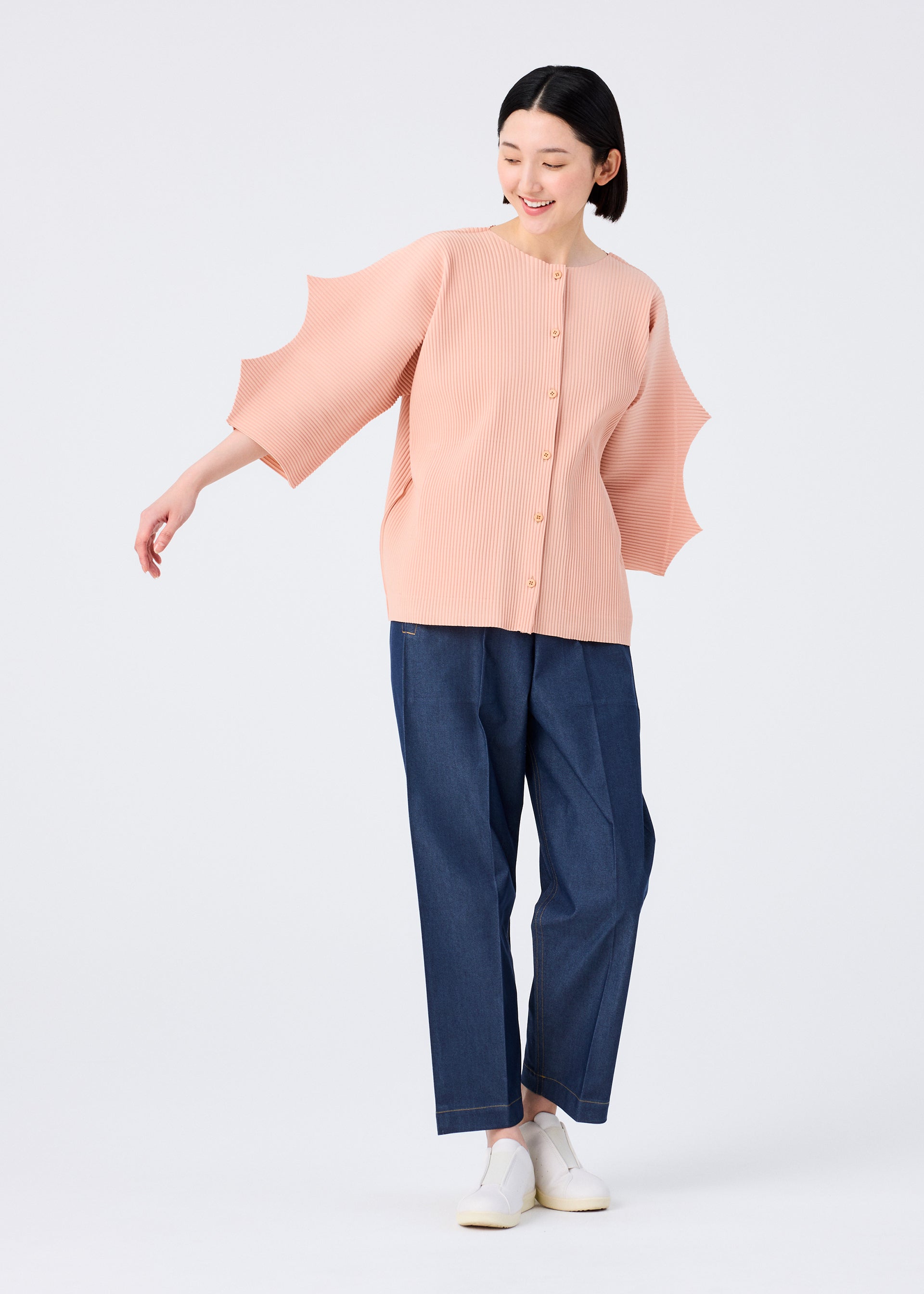Zoomed view A woman smiles while wearing the me ISSEY MIYAKE GIZA GIZA PLEATS peach knit cardigan with sculpted sleeves, paired with blue wide-leg pants and white slip-on shoes, standing against a plain light background.