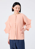 A woman with short black hair smiles gently, looking down with one hand in her pocket, wearing the me ISSEY MIYAKE GIZA GIZA PLEATS light pink pleated knit cardigan and dark blue pants against a plain light background.