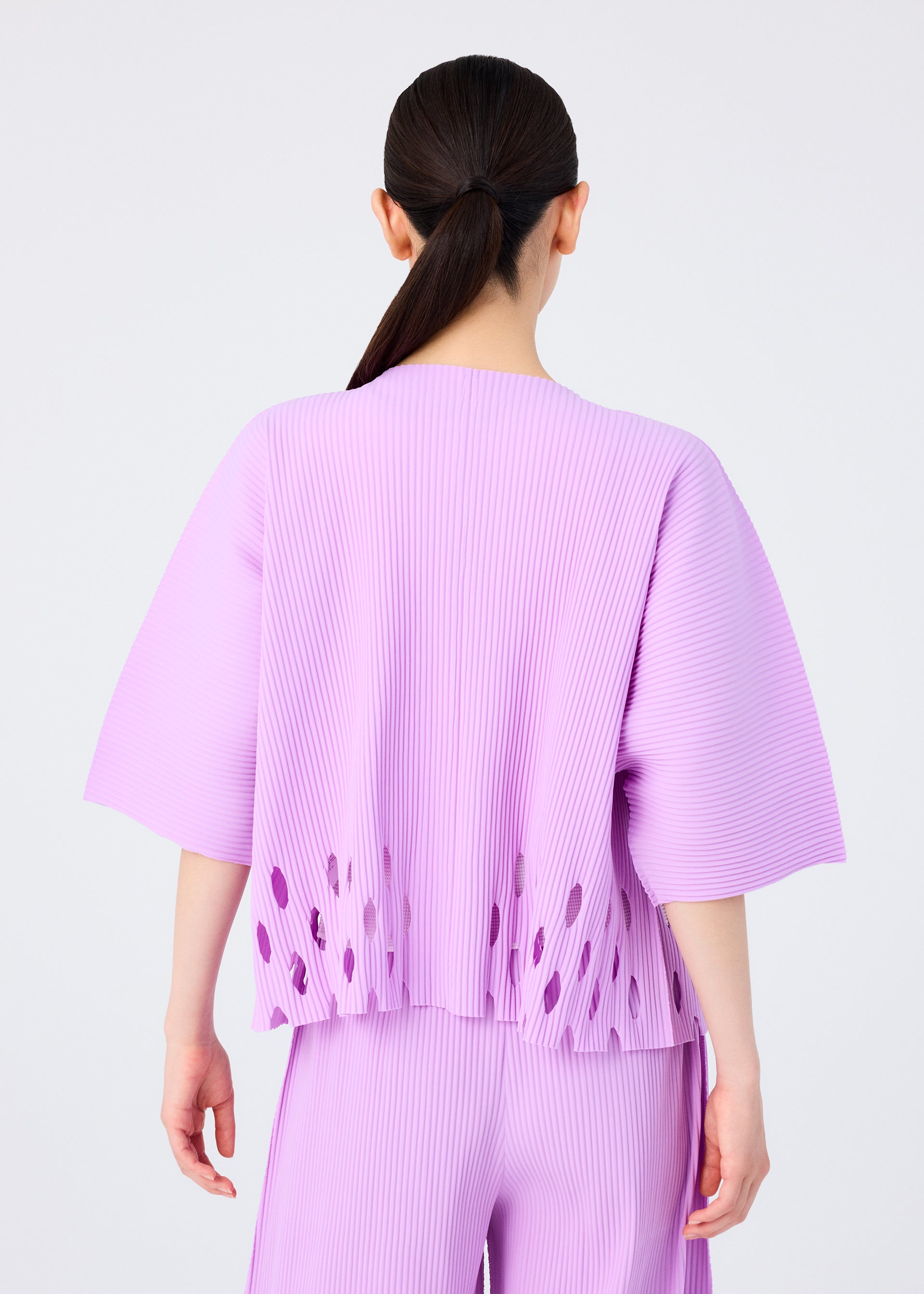 Zoomed view A person with long dark hair in a low ponytail is shown from the back, wearing the me ISSEY MIYAKE APERTURE PLEATS set—a loose-fitting, light purple pleated knit top with oval cut-outs near the hem and matching pants.