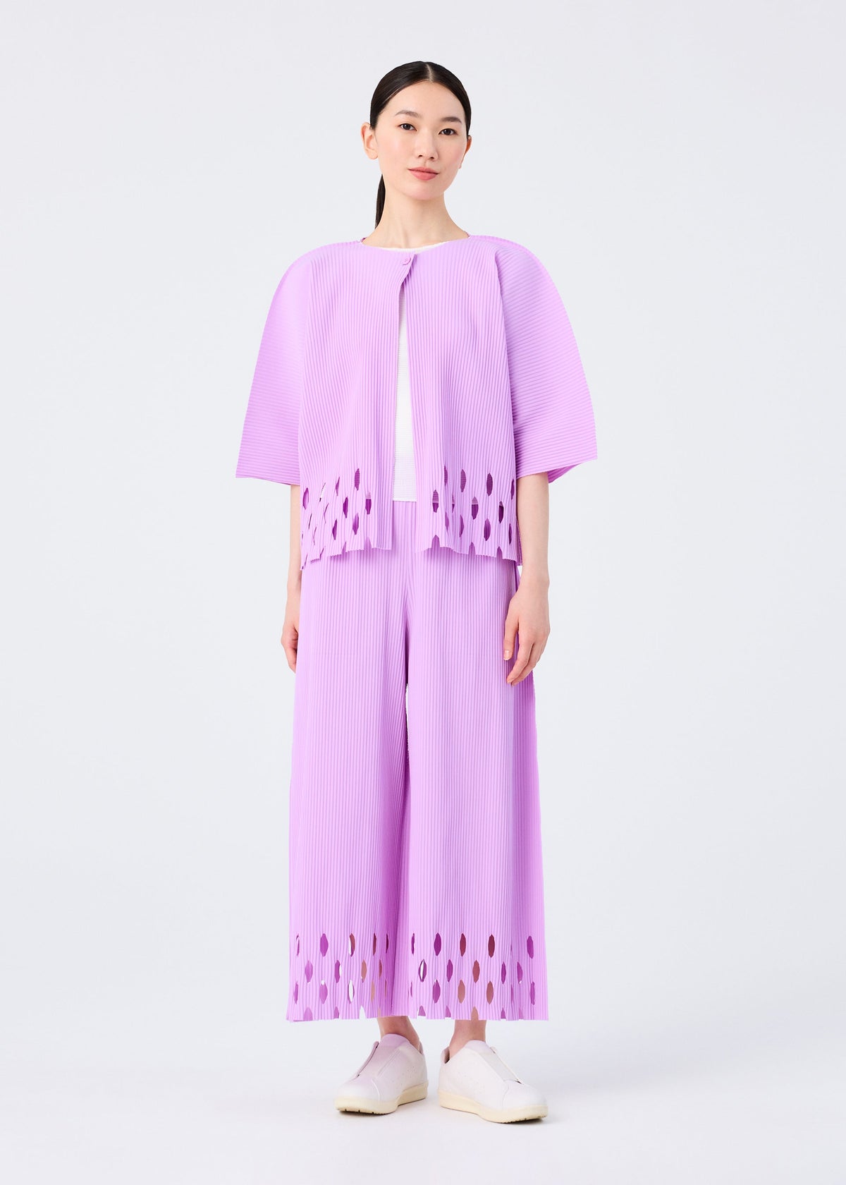 A woman with straight dark hair and white shoes wears the APERTURE PLEATS set by me ISSEY MIYAKE: a matching light purple pleated knit top and wide-leg pants with oval cut-out hems, standing against a plain white background. view 7 of 7