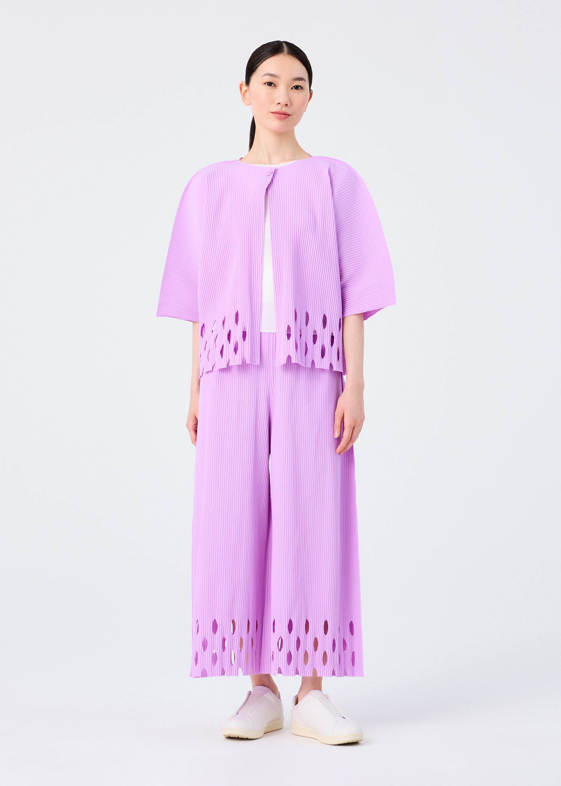 Zoomed view A woman with straight dark hair and white shoes wears the APERTURE PLEATS set by me ISSEY MIYAKE: a matching light purple pleated knit top and wide-leg pants with oval cut-out hems, standing against a plain white background.