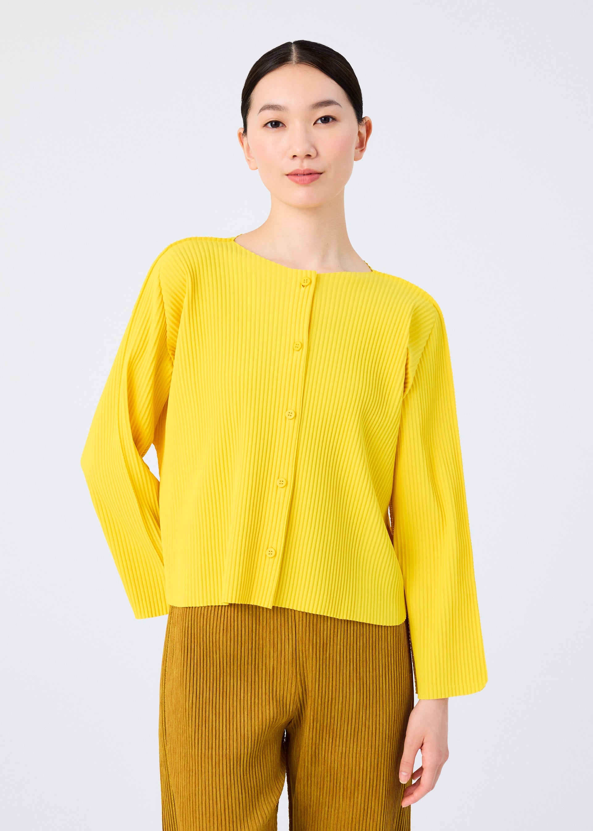 Zoomed view A person stands against a white background wearing the me ISSEY MIYAKE ROUND PLEATS: a bright yellow, long-sleeve pleated cardigan with vertical pleats, paired with light brown pleated pants.