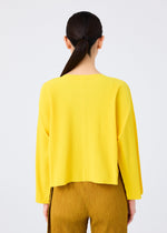 A woman with straight dark hair in a low ponytail is seen from behind, wearing me ISSEY MIYAKE ROUND PLEATS mustard-colored pleated pants and a loose cardigan against a plain white background.