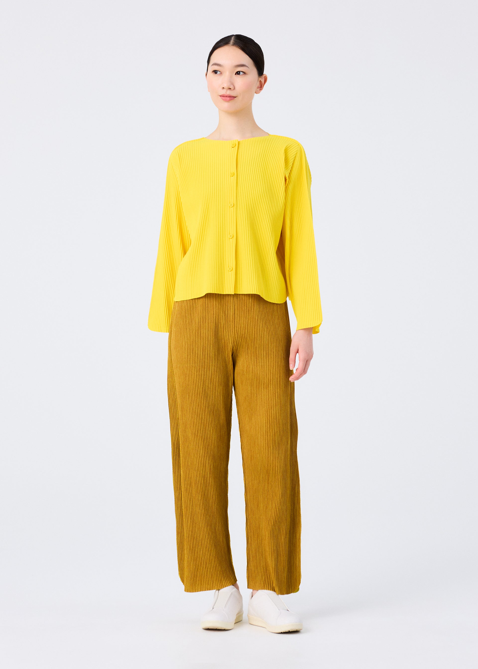 Zoomed view A person stands against a plain white background, wearing the me ISSEY MIYAKE ROUND PLEATS long-sleeve button-up yellow cardigan and loose-fitting mustard yellow pants, paired with white shoes.