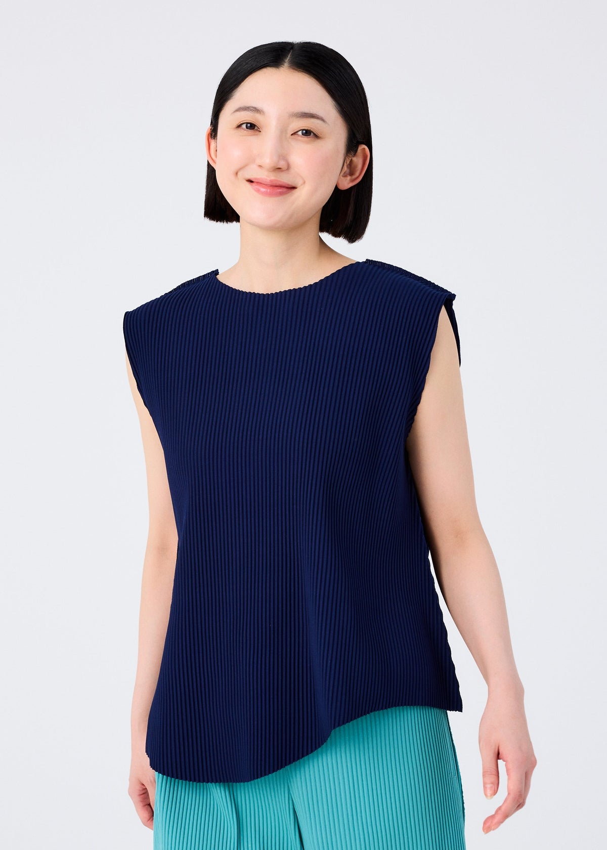 A woman with straight, chin-length dark hair smiles in a navy blue sleeveless top and teal pants from the AURORA PLEATS collection by me ISSEY MIYAKE, standing against a plain light background. view 6 of 8