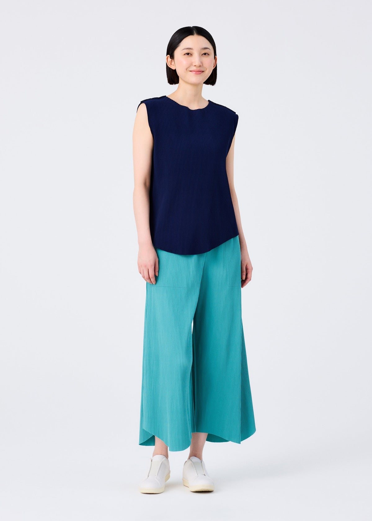A woman stands facing forward in the AURORA PLEATS sleeveless navy blue top by me ISSEY MIYAKE, loose teal wide-leg pants, and white slip-on shoes. She has straight shoulder-length dark hair and smiles slightly against a plain white background. view 8 of 8