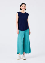 A woman stands facing forward in the AURORA PLEATS sleeveless navy blue top by me ISSEY MIYAKE, loose teal wide-leg pants, and white slip-on shoes. She has straight shoulder-length dark hair and smiles slightly against a plain white background.