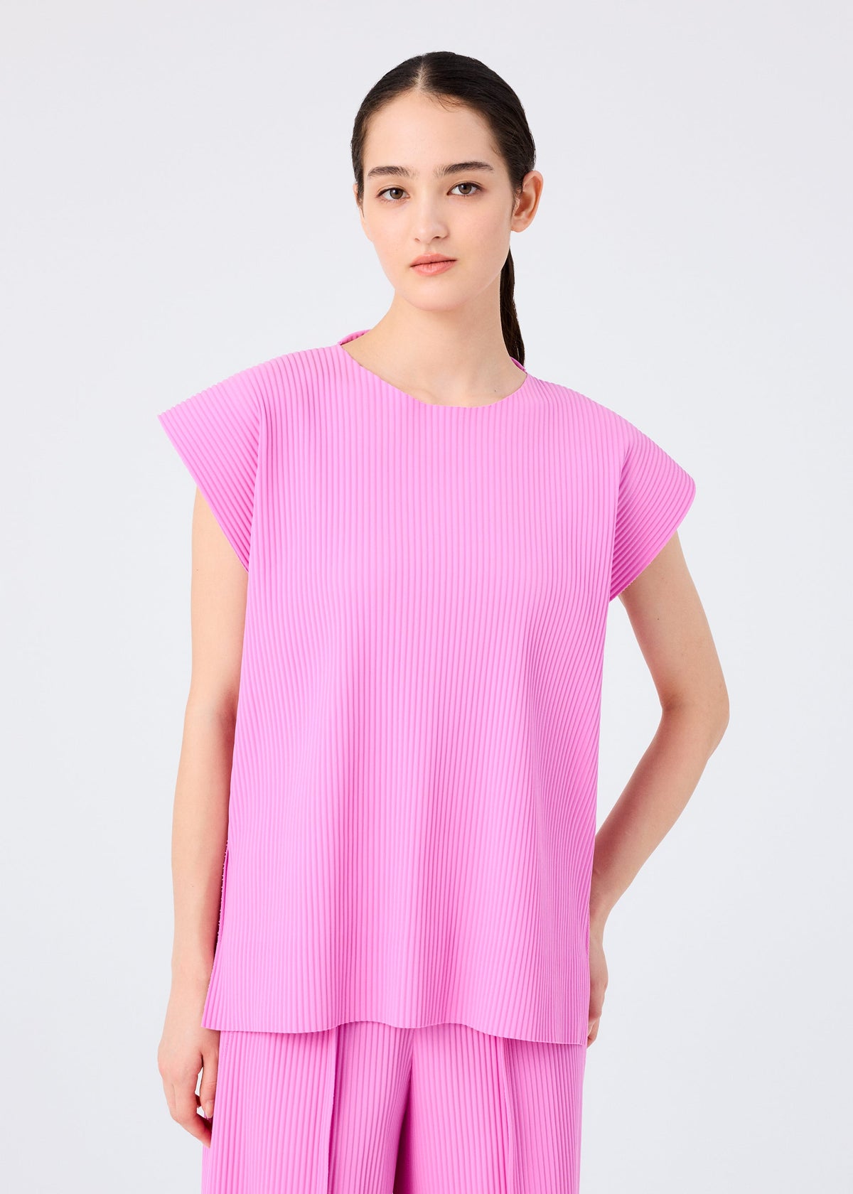 A woman with straight, dark hair pulled back wears the me ISSEY MIYAKE TRICK PLEATS set— a loose-fitting, fine knit pleats top with French sleeves and matching wide-leg pants—against a plain background, her expression neutral. view 6 of 8