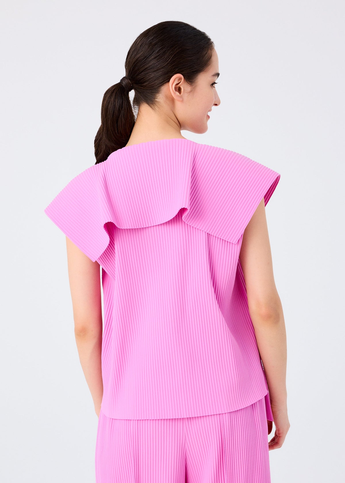 A woman with dark hair in a ponytail is seen from behind wearing the bright pink TRICK PLEATS top and pleated pants by me ISSEY MIYAKE, featuring a large ruffled collar. The background is plain and light-colored. view 7 of 8