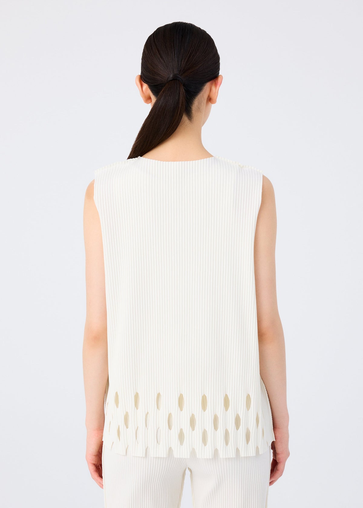 A woman with long dark hair in a low ponytail is seen from behind, wearing the APERTURE PLEATS sleeveless top by me ISSEY MIYAKE, featuring oval cut-outs and paired with matching white pants against a plain background. view 6 of 7