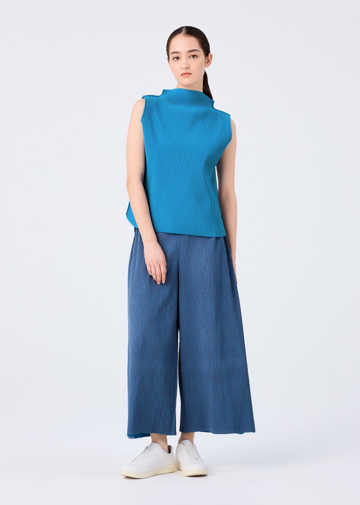 A woman with long, straight hair pulled back stands against a plain background, wearing the ROUND PLEATS sleeveless blue knit top and wide-leg pants from me ISSEY MIYAKE, paired with white slip-on shoes and a neutral expression. view 7 of 7