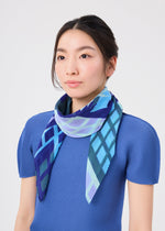 A woman with straight dark hair wears a blue textured top and the BUILDING PLEATS STOLE by me ISSEY MIYAKE, which features a blue geometric pattern, standing against a plain white background.