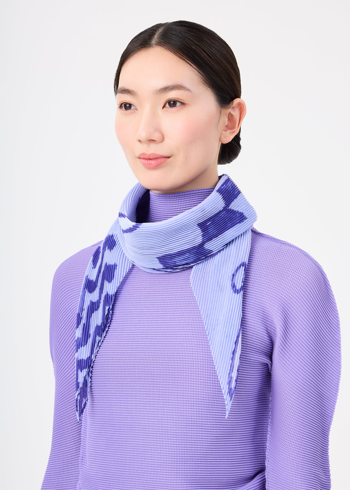 A woman with straight dark hair in a low bun stands against a plain white background, wearing a lavender textured top and the me ISSEY MIYAKE PLANT PLEATS STOLE with a plant pattern draped around her neck. view 6 of 6