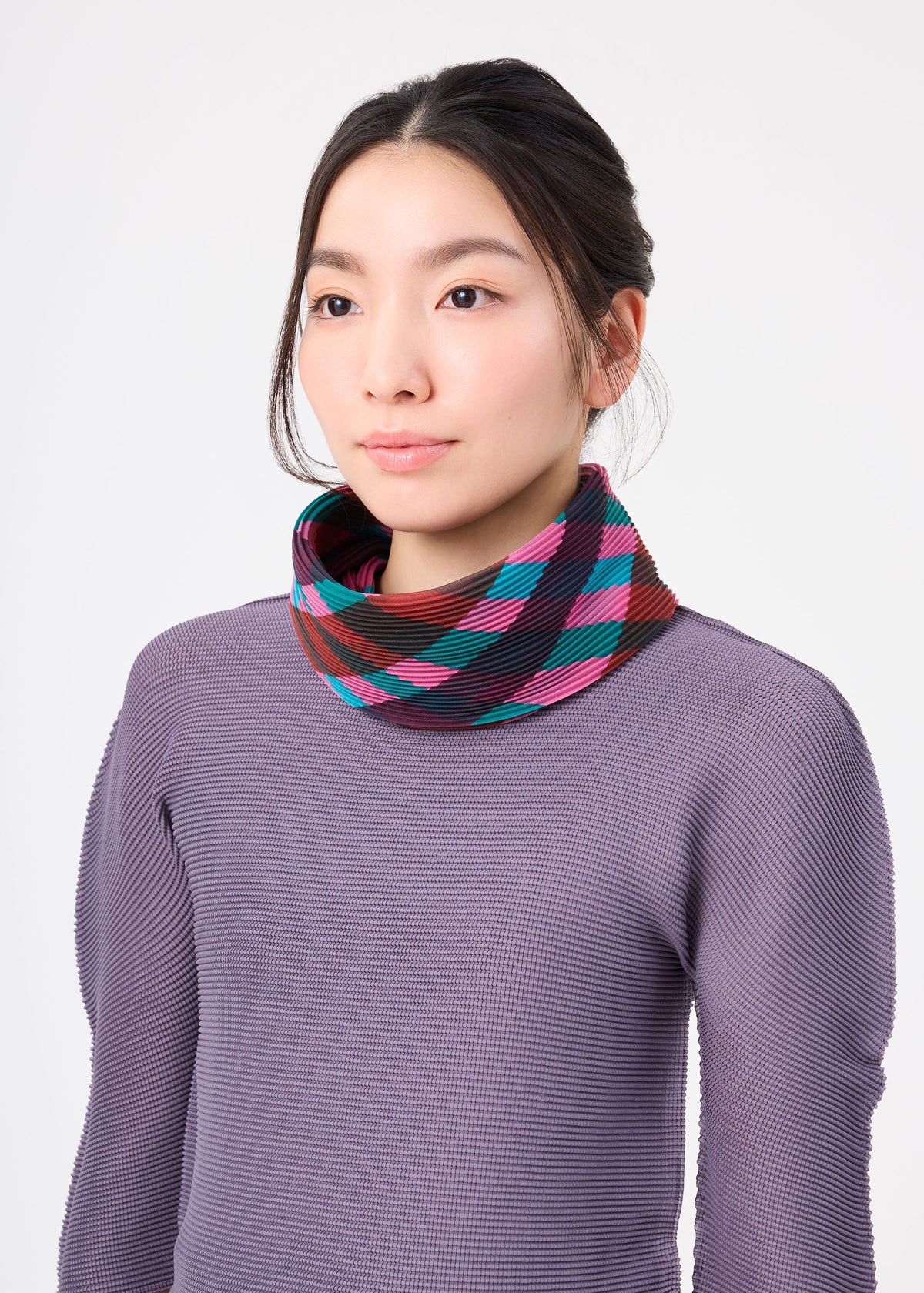 Zoomed view A woman in a textured purple top and the TAPE CHECK PLEATS STOLE by me ISSEY MIYAKE, featuring colorful stripes, stands against a white background, gazing slightly to the side with a neutral expression.