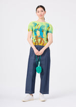 A person stands facing forward against a plain white background, holding the me ISSEY MIYAKE FRUITS POUCH and a green pom-pom ribbon accessory, while wearing a colorful tropical print shirt, wide-leg blue pants, and white shoes.