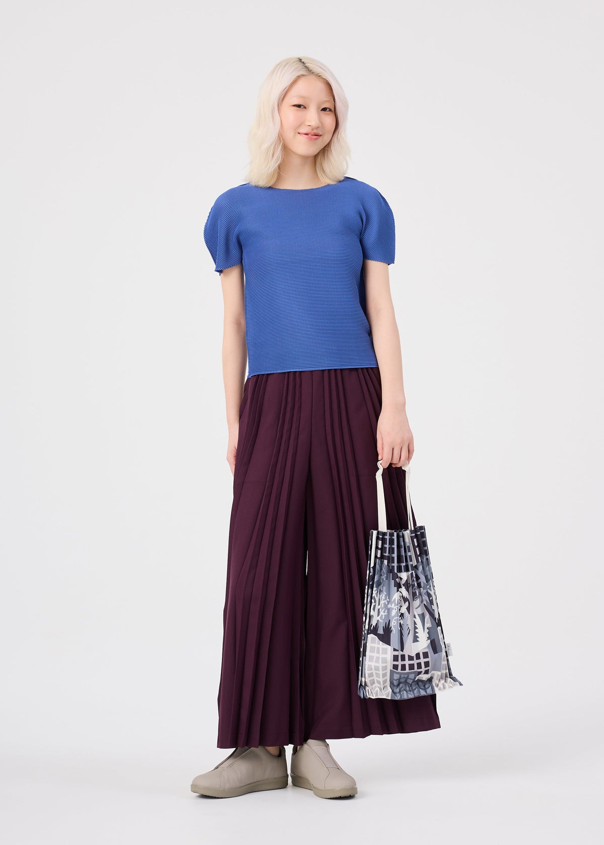 A person with light hair stands against a plain white background, dressed in a blue top, wide burgundy pleated pants, and beige shoes, while holding the me ISSEY MIYAKE BUILDING TRUNK PLEATS BAG featuring a printed pattern. view 8 of 8
