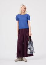 A person with light hair stands against a plain white background, dressed in a blue top, wide burgundy pleated pants, and beige shoes, while holding the me ISSEY MIYAKE BUILDING TRUNK PLEATS BAG featuring a printed pattern.