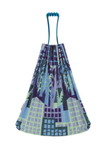 The BUILDING TRUNK PLEATS BAG by me ISSEY MIYAKE is a reusable tote featuring blue handles and an artistic cityscape pattern in shades of blue, purple, and green, with buildings, trees, and subtle urban farming elements.