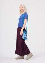 A person with light hair wearing a blue top, long burgundy skirt, tan shoes, and carrying the me ISSEY MIYAKE BUILDING TRUNK PLEATS BAG with its distinctive handle over their shoulder stands against a plain white background.
